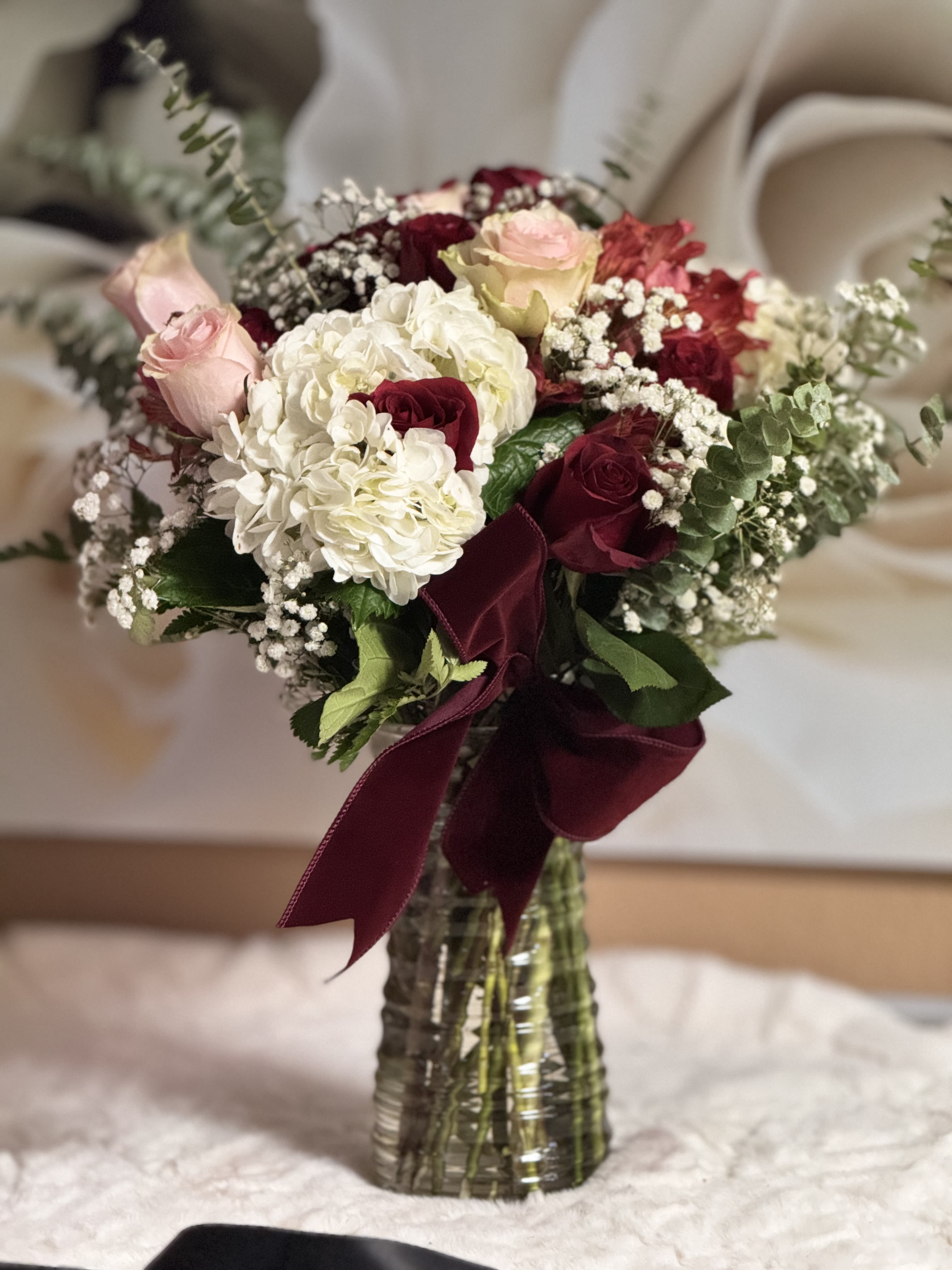 Tall vase arrangement of red and pink roses with white hydrangeas and baby's breath, tied with a burgundy bow.