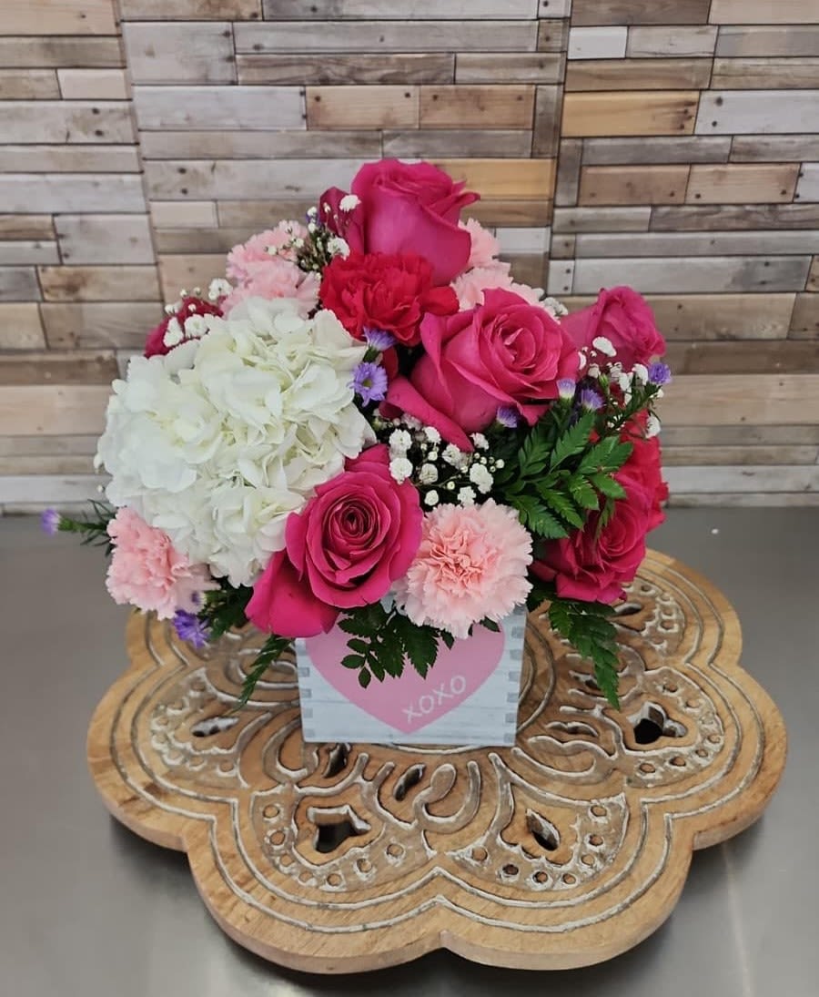 Compact arrangement of hot pink roses, white hydrangea, and pink carnations in a heart-print cube container.