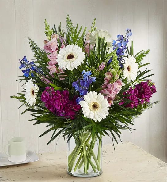 Mixed bouquet of white gerbera daisies, pink blooms, and blue flowers in a glass vase