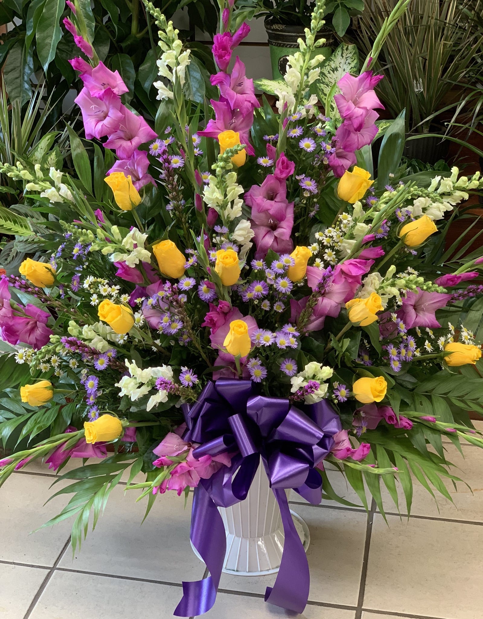 Large mixed floral arrangement in a white pedestal vase with a purple bow