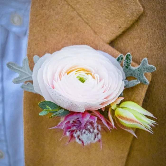 White ranunculus boutonniere with pink and yellow accents on a tan jacket