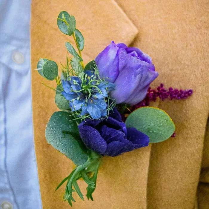 Purple boutonniere with blue flower and eucalyptus on tan wrap