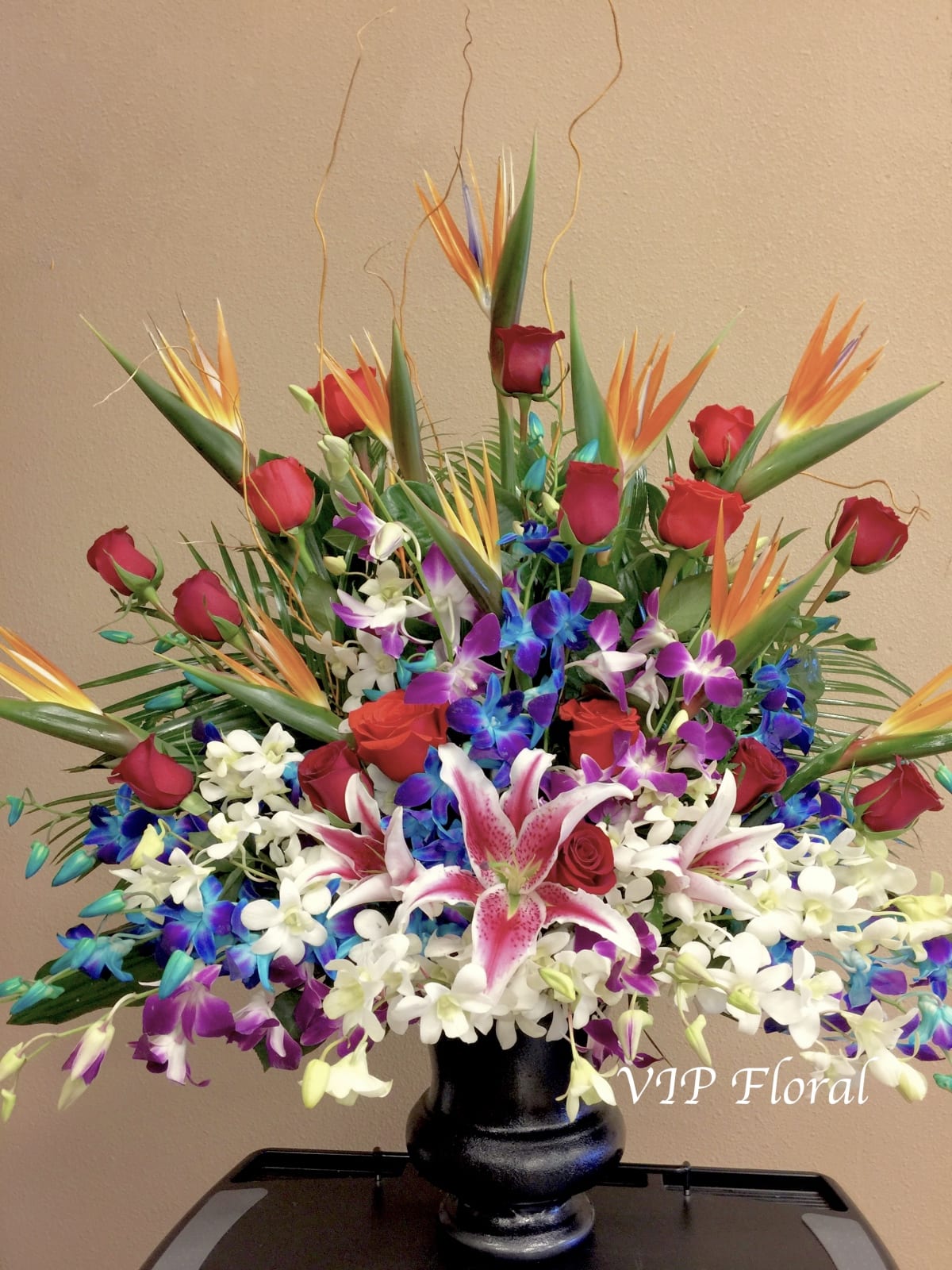 Large tropical bouquet with red roses, orchids, and bird of paradise in a black vase