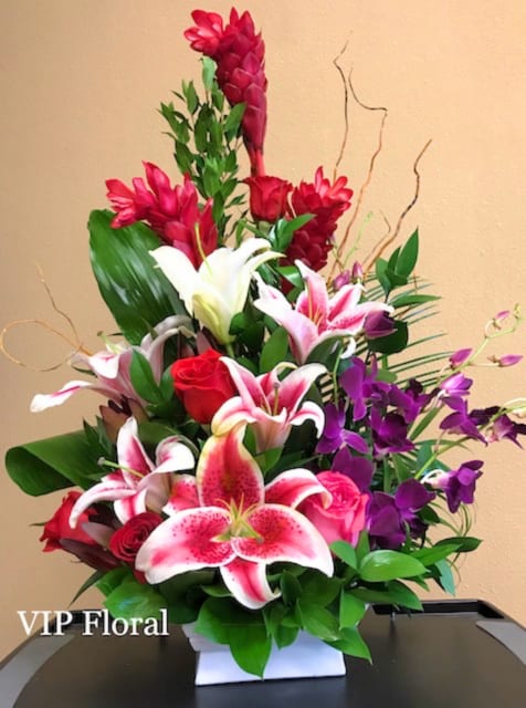 Tall floral arrangement with pink lilies, red roses, and purple orchids in a white container