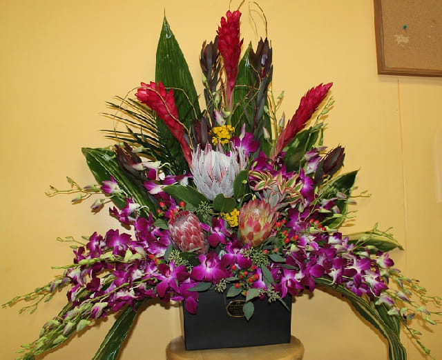 Large tropical flower arrangement in a black container with purple orchids and red blooms