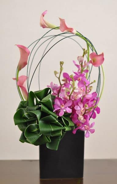 Pink calla lilies and orchids in a black vase with arched stems