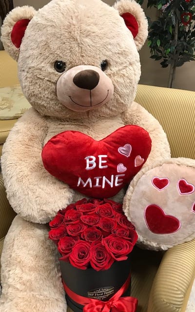 Large teddy bear holding a red heart pillow and a box of red roses
