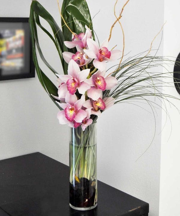 Pink orchids arranged in a tall glass vase with long decorative leaves