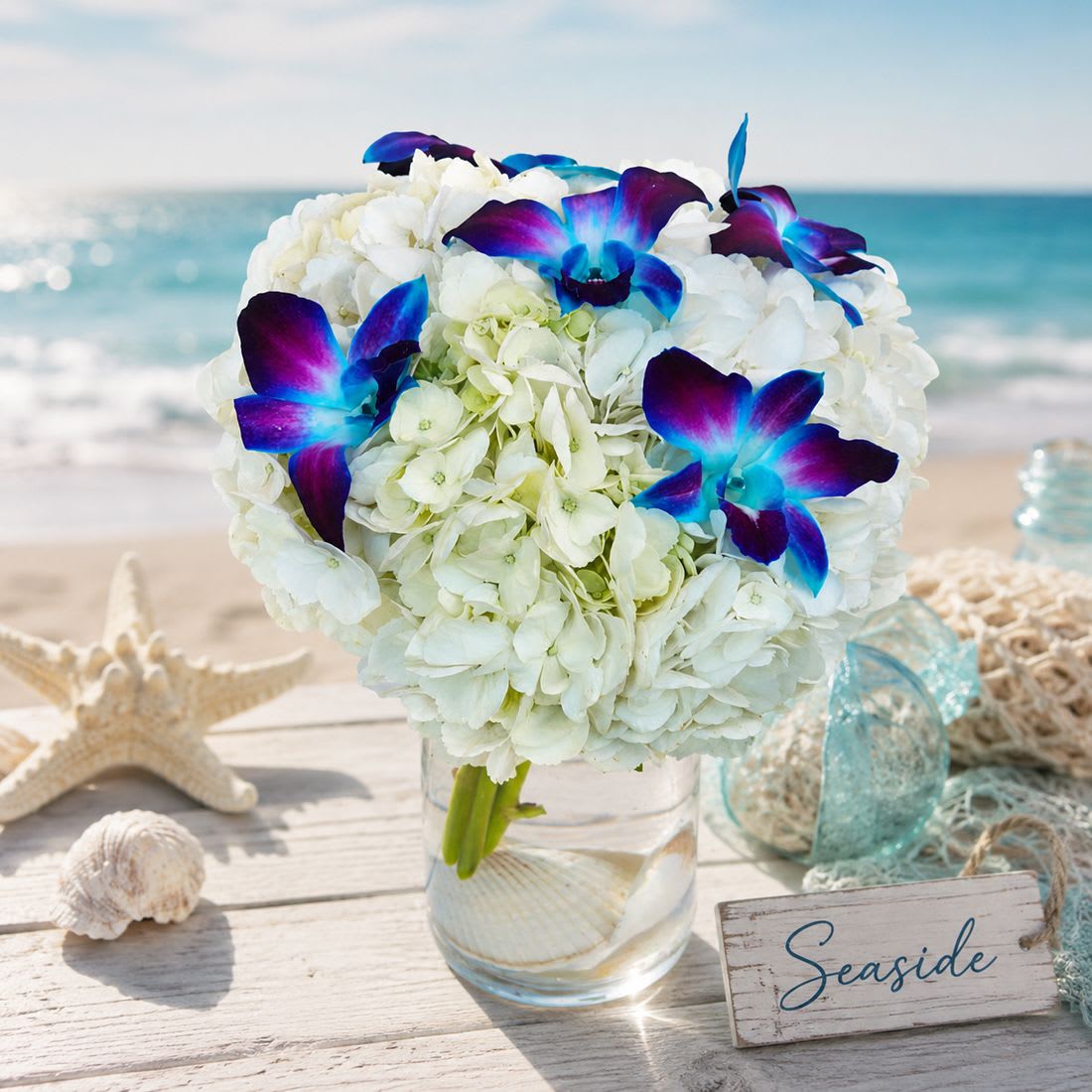 Seaside - Seaside Floral Arrangement – Hydrangeas &amp; Blue Orchids  Bring the calming beauty of the ocean into any space with our Seaside arrangement, featuring lush white hydrangeas and exotic blue dendrobium orchids resting on a bed of seashells. Designed in a sleek clear glass cylinder vase, this coastal-inspired bouquet makes a stunning gift for birthdays, anniversaries, or simply to brighten someone’s day.  Available Sizes:  • Standard – 3 white hydrangeas &amp; 5 blue dendrobium orchid blooms in a 4" cylinder vase (approx. 8" W x 12" H)  • Deluxe – 4 white hydrangeas &amp; 8 blue dendrobium orchid blooms in a 4" cylinder vase  • Premium – 6 white hydrangeas &amp; 12 blue dendrobium orchid blooms in an upgraded 6" cylinder vase  Bring a touch of seaside serenity to their home with this hydrangea and orchid flower arrangement—a gift as refreshing as the ocean breeze.