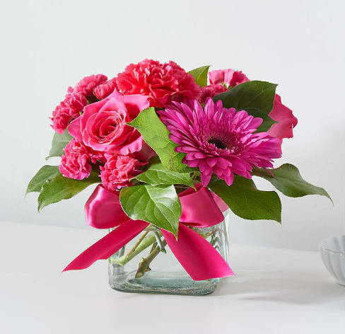 Bright pink roses, carnations, and a gerbera daisy in a square glass vase with a pink ribbon bow.