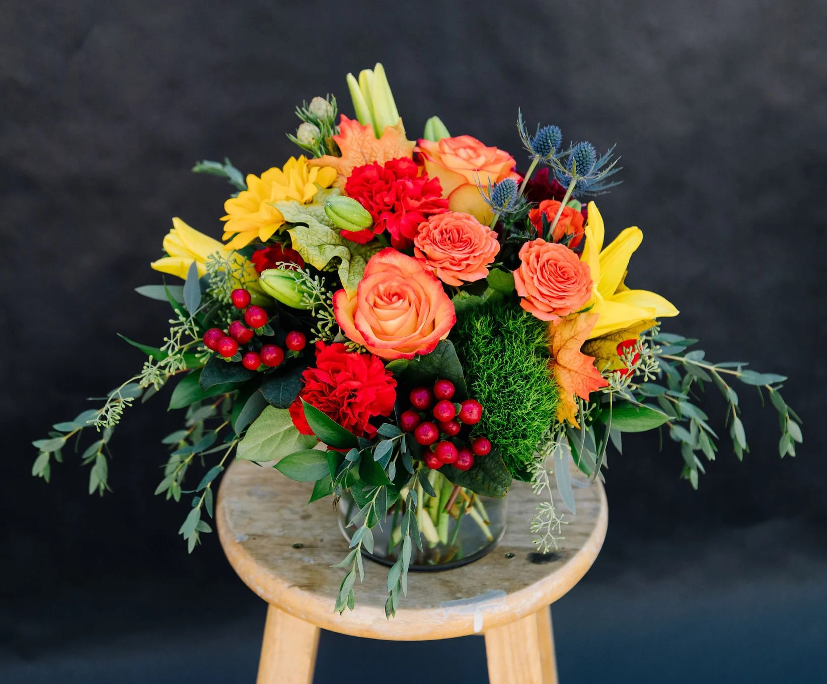 Warm Fall Wishes - Celebrate the beauty of Fall with this lush, colorful heartwarming mix of roses, lilies sunflowers and carnations designed and delivered in a low cylinder vase.  Accents of eucalyptus, fall leaves and hypericum berries add the final touch.