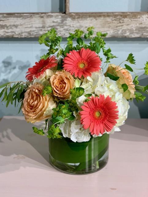 Short vase arrangement with coral gerbera daisies, peach roses, and white hydrangeas in a clear glass cylinder