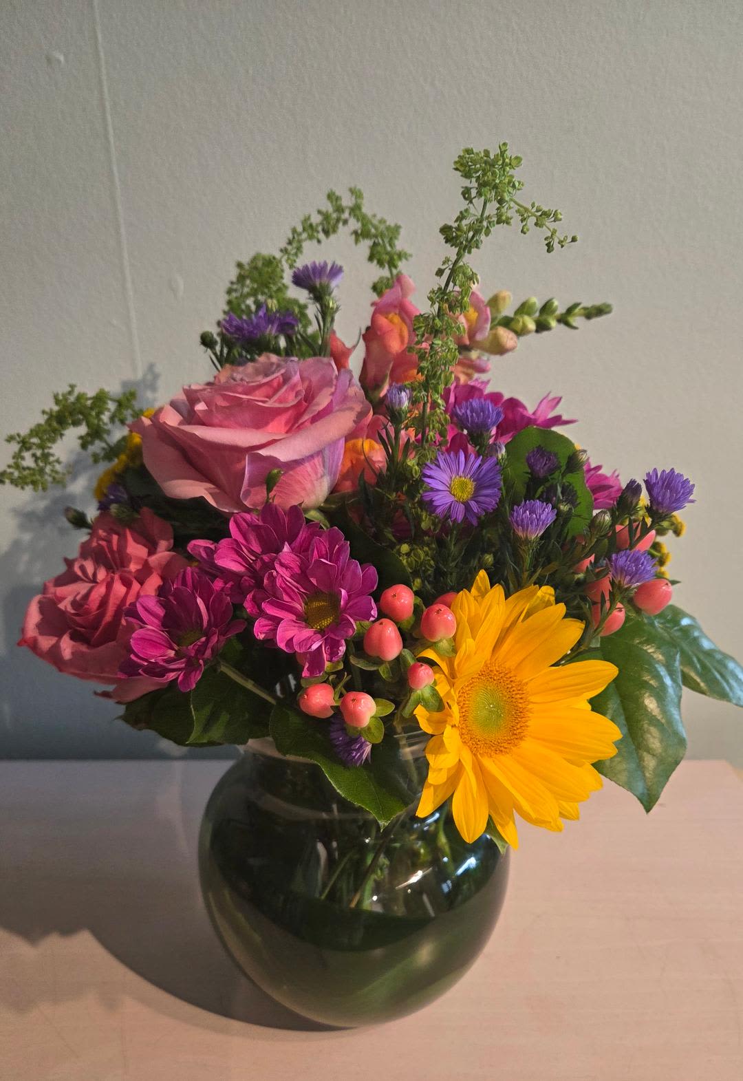 Mixed bouquet of pink roses, purple daisies, and a yellow gerbera in a glass vase