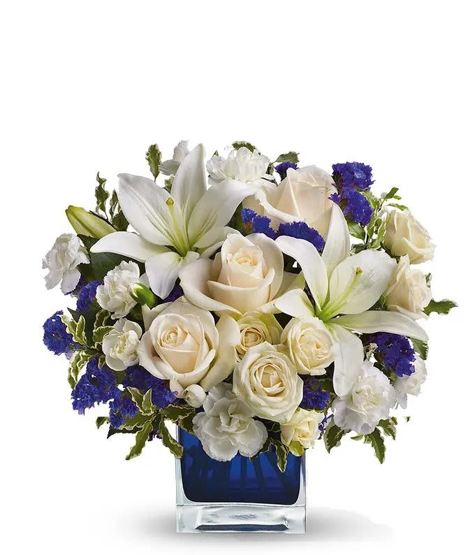 Sapphire Skies Bouquet - Send someone a bit of heaven with this beautiful bouquet. Luxurious crème roses and pure white lilies paint a peaceful picture inside a sapphire blue cube.  Crème roses, white asiatic lilies and white miniature carnations are mixed with bursts of purple statice and green pitta negra. Delivered in a glass Cube.  Approximately 12" W x 11 1/2" H  Orientation: All-Around  As Shown : TEV25-3A Deluxe : TEV25-3B Premium : TEV25-3C 
