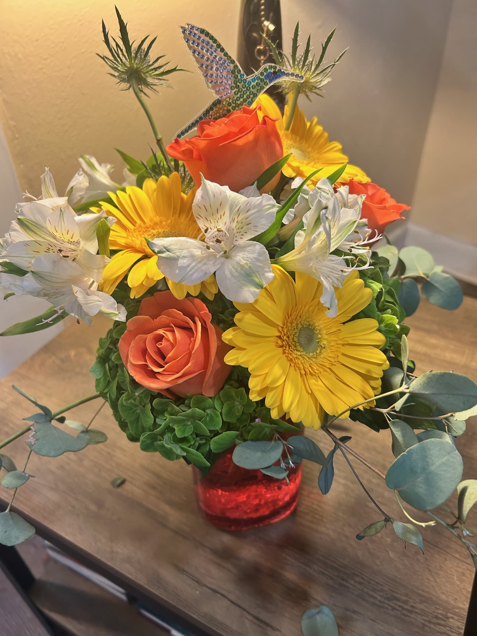 Summer Sorbet - Sunlit orange roses and bright yellow gerbera daisies make this flower arrangement a joyful birthday bouquet or thank-you gift that instantly lifts the mood. White alstroemeria and cool eucalyptus foliage keep the warm palette fresh and balanced, while a jeweled hummingbird pick adds a playful, celebratory touch your recipient will notice.  Arranged compactly in a red glass vase, the rounded design reads cheerful but casual-perfect for spring occasions, a hostess gift, or "just because" surprises. Colors: yellow, orange, white, and green. Includes orange roses, yellow gerbera daisies, white alstroemeria, eucalyptus, a red glass vase, and a decorative hummingbird pick. The size and full, rounded shape make it easy to place on a table, desk, or entryway where it will brighten the room and make someone feel truly seen.