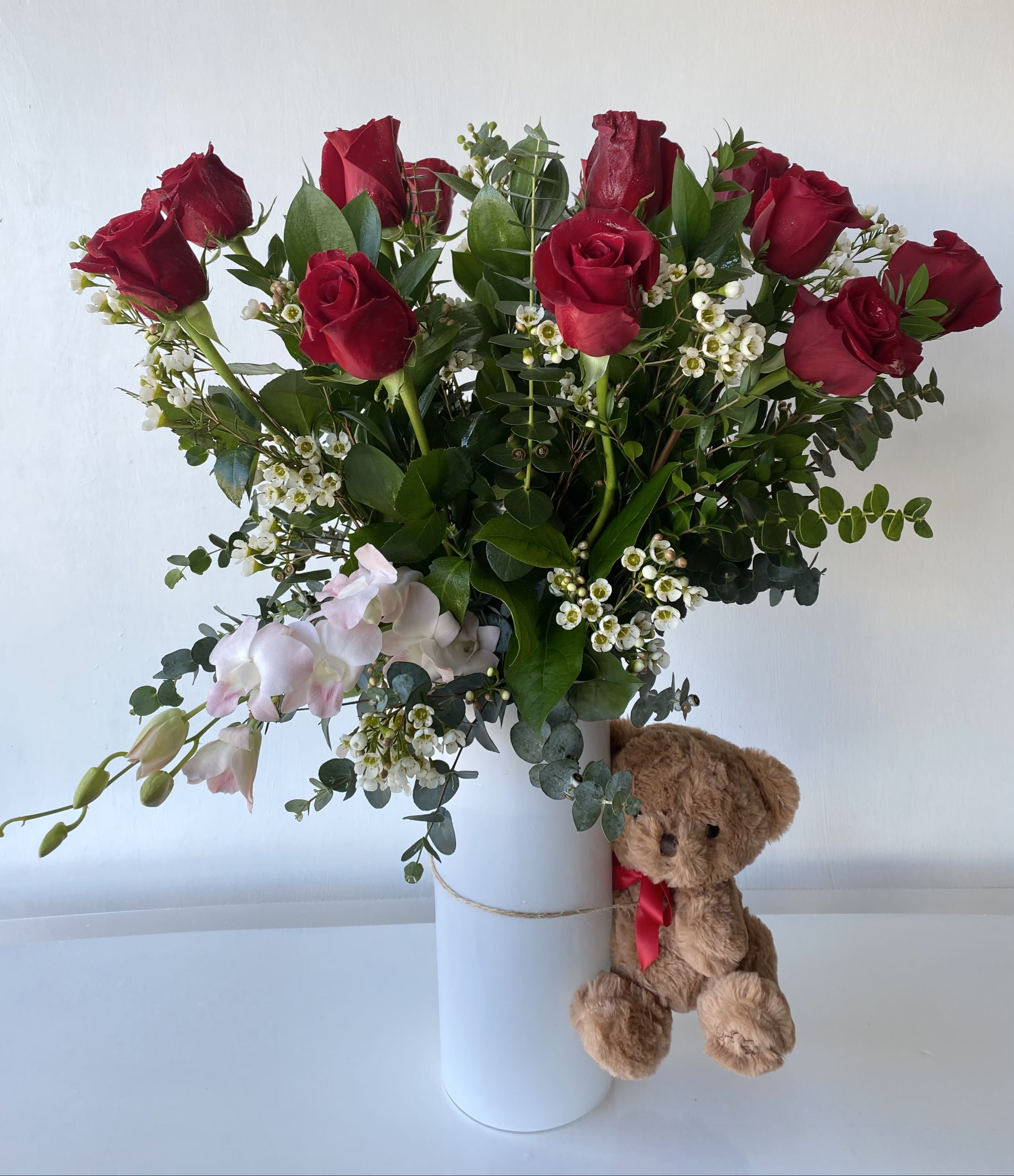My Valentine - This arrangement features 12 long stems of red roses with a branch of light pink dendrobium orchid. The arrangement in a 12" white glass cylinder vase with a brown teddy bear hugging it by the side. This arrangement is a cute Valentine's Day gift for your love.