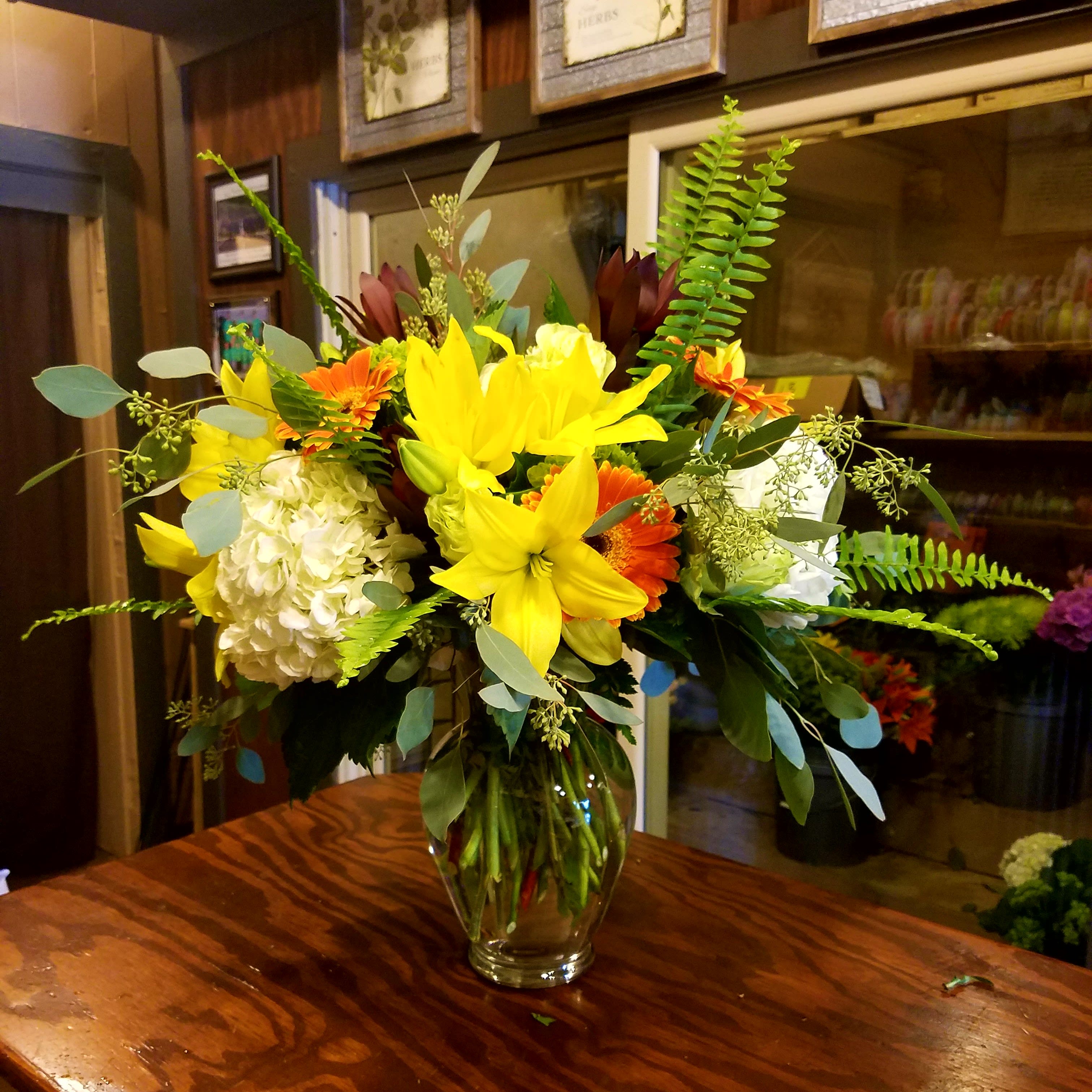 Mixed bouquet of yellow lilies, orange gerberas, white hydrangea, and greenery in a glass vase