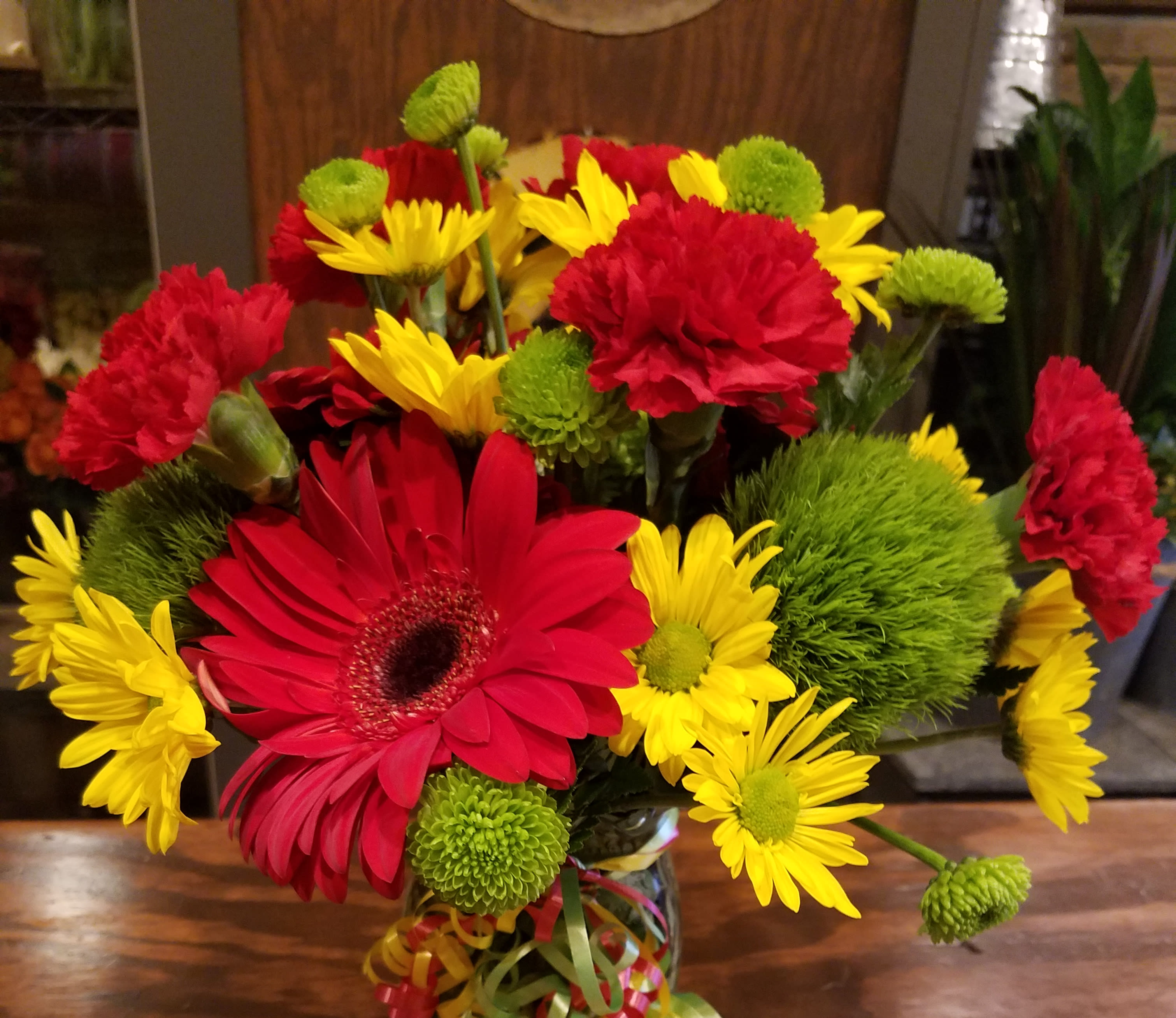 Bouquet of red gerbera daisies, yellow daisies, and green pompons in a glass vase