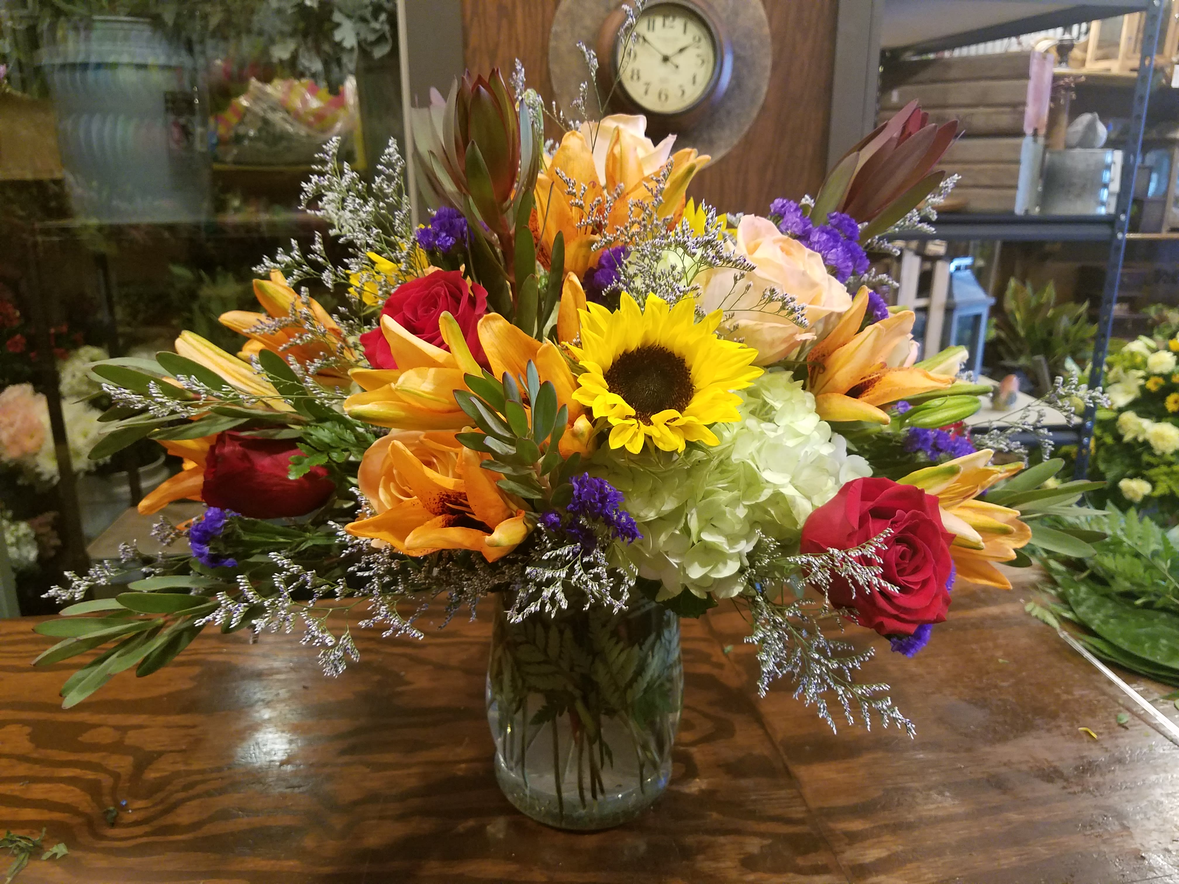 Mixed bouquet with sunflower, orange lilies, red roses, and hydrangea in a clear glass vase