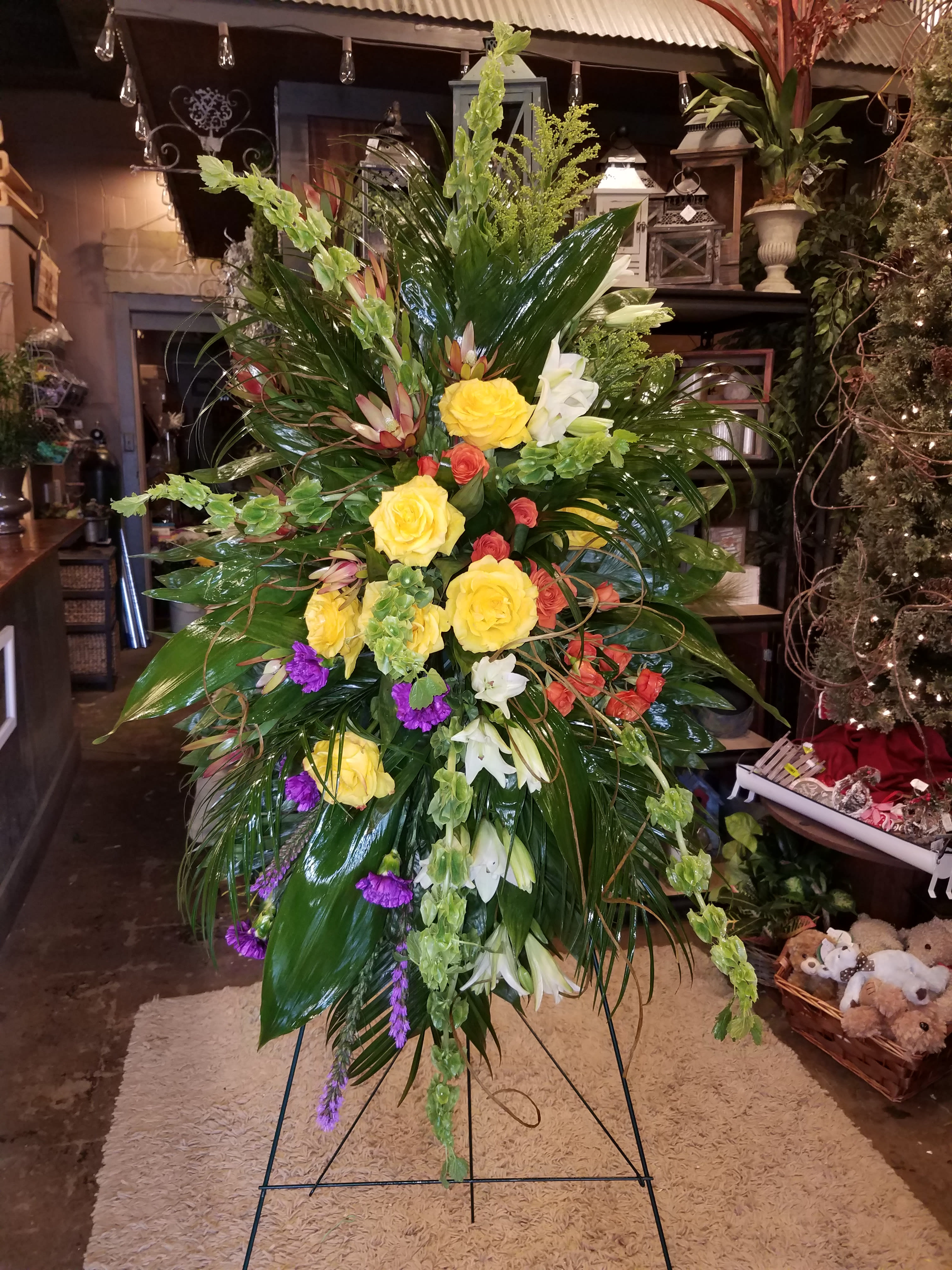Color block spray - Spray on a stand with a variety of flowers placed in a unique design. Some of the flowers included are, yellow roses, bells of irelsnd, and orange spray Rose's. 