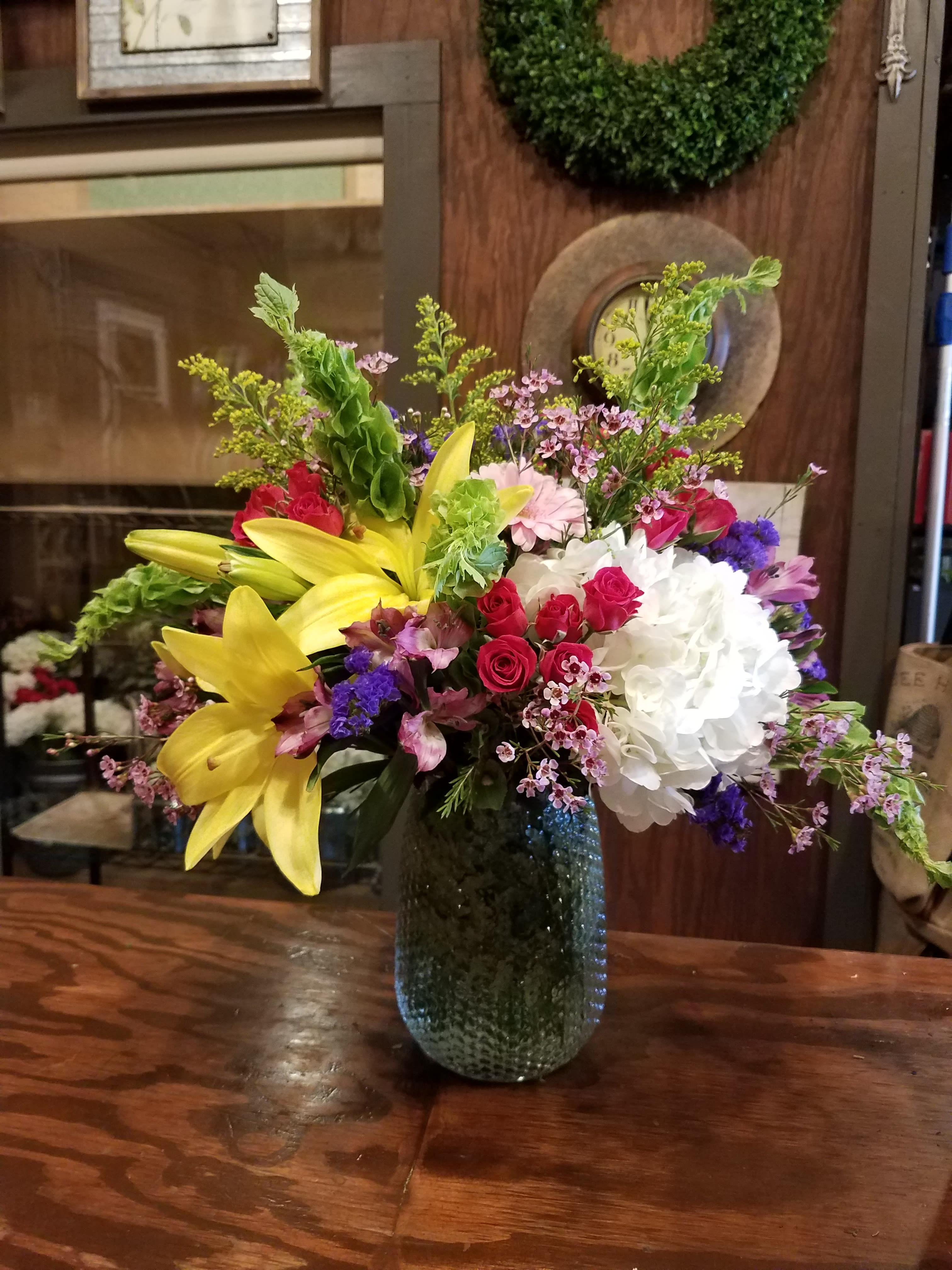 Mixed bouquet with yellow lilies, red roses, white hydrangea, and purple accents in a glass vase