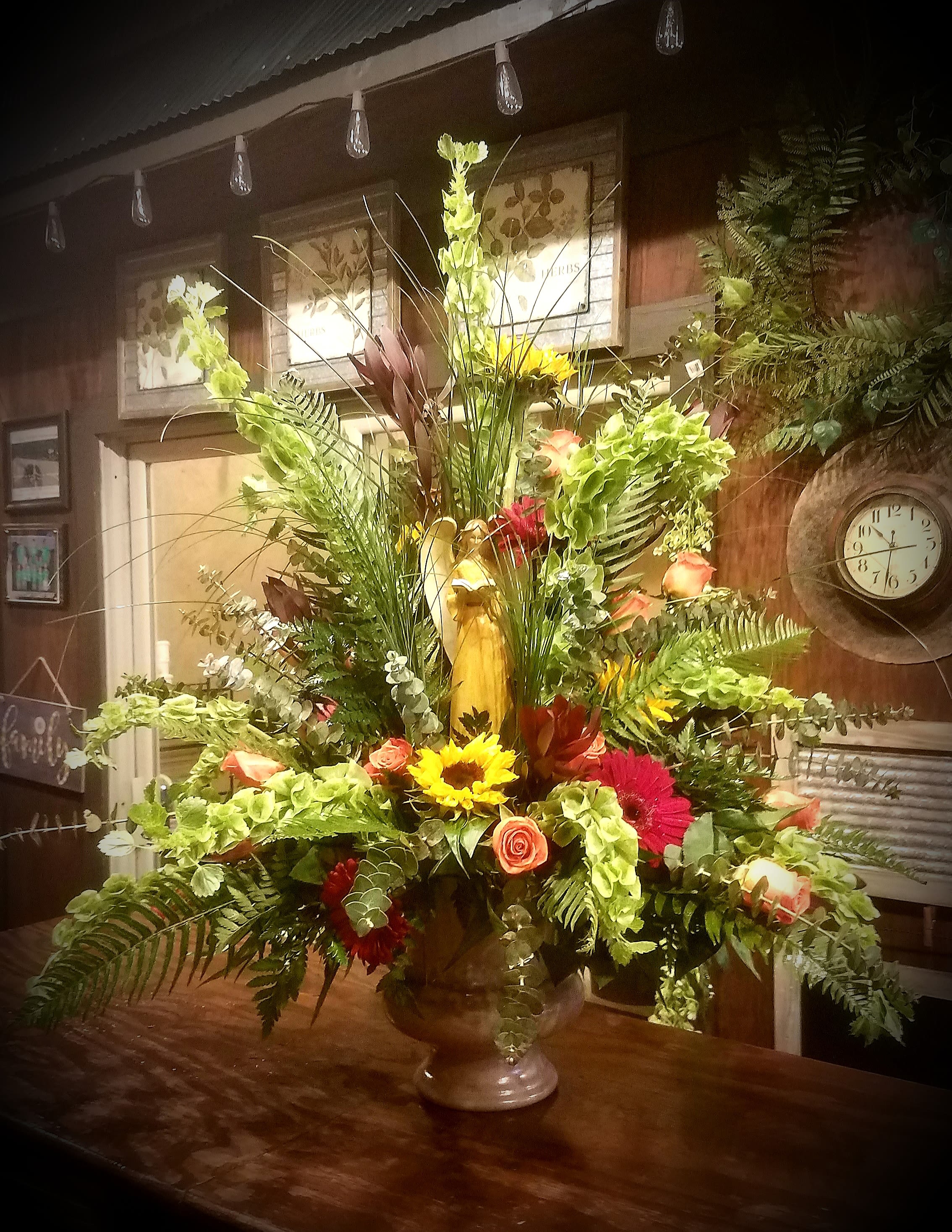 Angel Sympathy basket - This large (3.5' × 3.5') urn arrangement features an Angel. It can be custom ordered to color. This arrangement features, bells ofIreland, sunflowers, Rose's, gerbera daisys , safari sunset, protea, and various filler. 