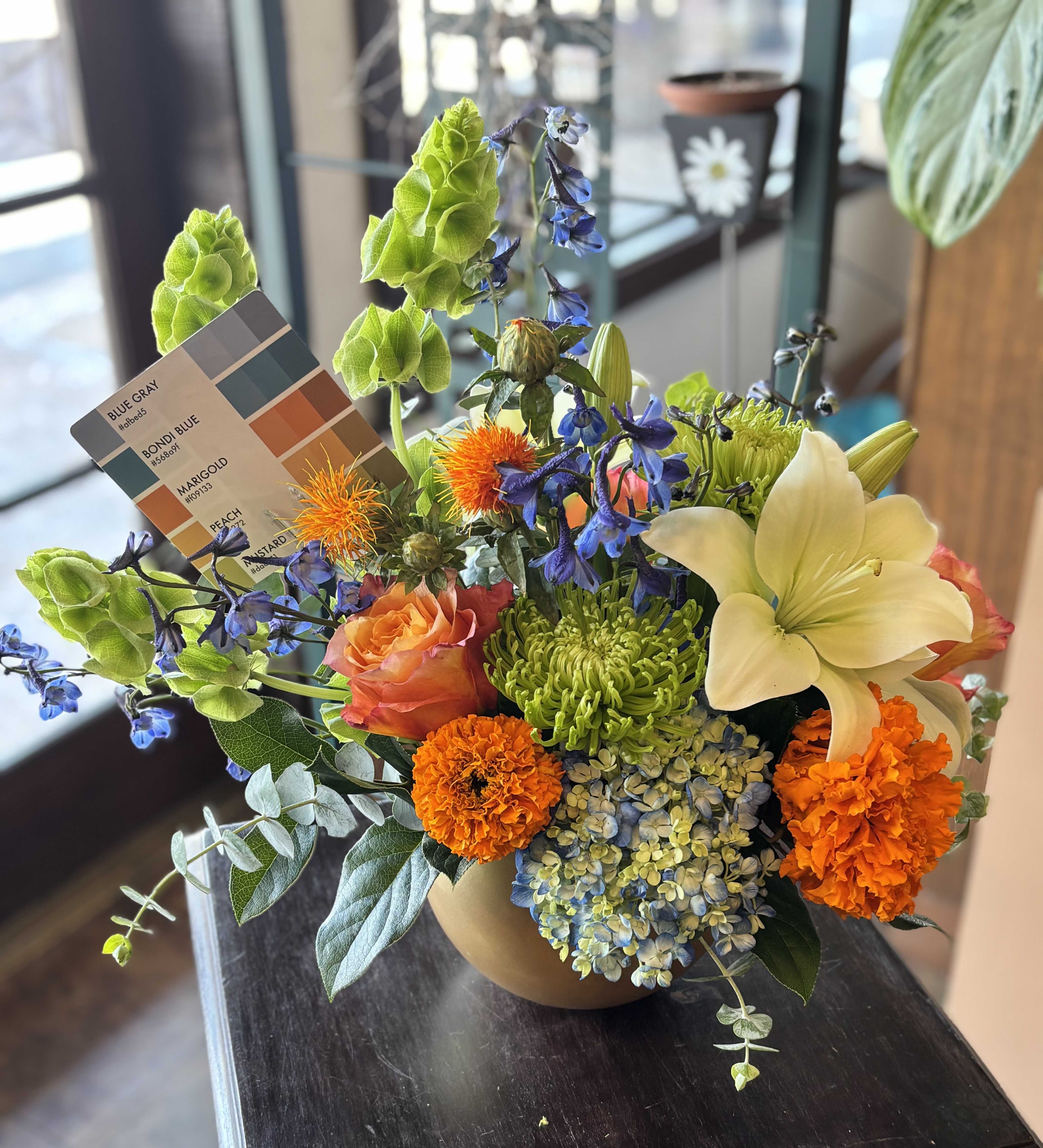 Mixed bouquet in a tan vase with orange, blue, green, and cream flowers.
