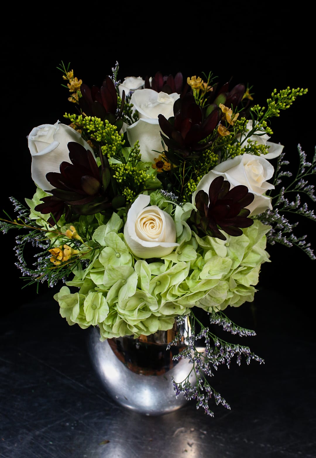 White roses and green hydrangeas in a silver vase with dark accent flowers