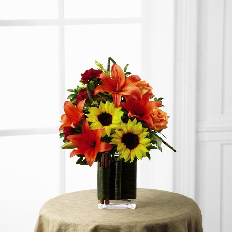 FTD Vibrant Views Bouquet - The FTD Vibrant Views Bouquet is a cheerful display of autumn delight to bring warm cheer to your special recipient with each sunlit bloom. Gorgeous sunflowers bring their beauty to this arrangement along with orange roses, deep orange Asiatic lilies, burgundy carnations, yellow solidago, and a variety of lush greens. Arriving in a clear rectangular glass vase wrapped with a green leaf material secured with cinnamon sticks for a sophisticated and modern look, this bouquet is a rare fall treat. 