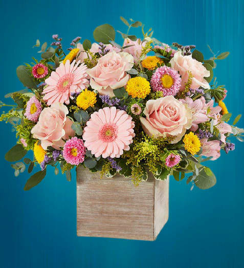 Spring Sentiment Bouquet - Spring is a time to refresh and reach out. Our new spring bouquet celebrates that sentiment. Soft pink and yellow blooms are loosely gathered with lush greenery for style and texture. Designed in our rustic, grey-washed wooden cube, it’s a gift that delivers on your feelings in the most beautiful way. Arrangement with pink roses, mini Gerbera daisies, Matsumoto asters and Peruvian lilies (alstroemeria); yellow button poms and purple limonium; accented with assorted greenery • Artistically designed in our rustic, natural wood-grain cube