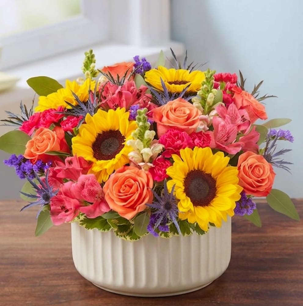 Bespoke Hatbox (Now in a Ceramic container!)  - EXCLUSIVE An exquisite gift with beautifully bespoke style. Our vibrant gathering of blooms is arranged in our signature European hatbox: an elegant, new dove gray container finished with a matching ribbon. Its versatile design is perfect for displaying a variety of colors, adding to the custom feeling of this special keepsake.  All-around arrangement with orange roses, sunflowers, pink snapdragons, hot pink Peruvian lilies (alstroemeria) and mini carnations, blue thistle and purple statice; accented with silver dollar eucalyptus and assorted greenery
