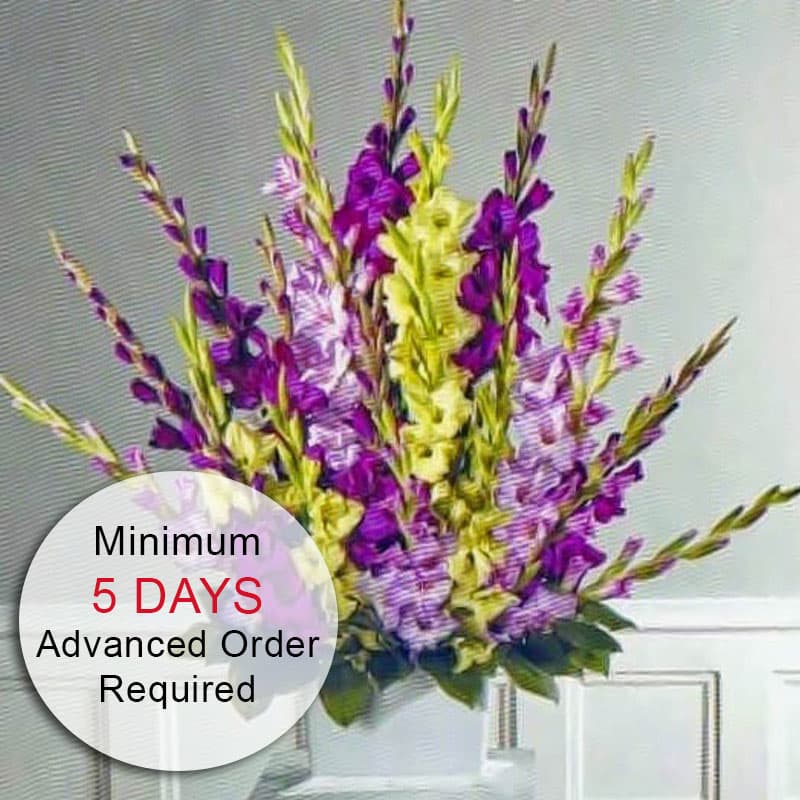 Tall fan-shaped arrangement of purple and pale yellow flowers in a white pedestal vase