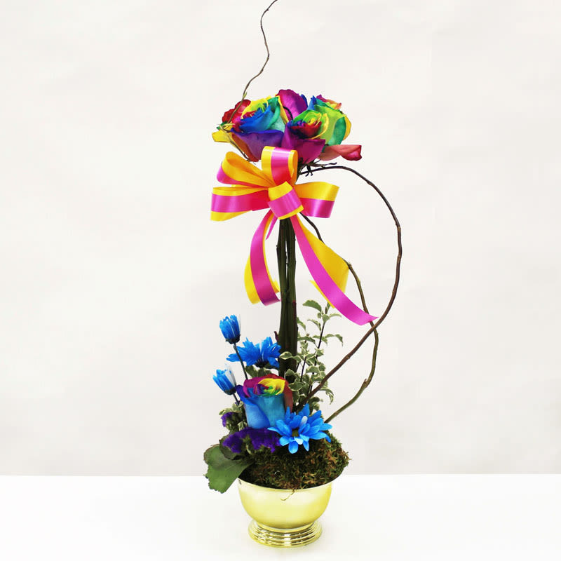 Tall arrangement of rainbow roses and blue flowers in a gold bowl with pink and yellow bow