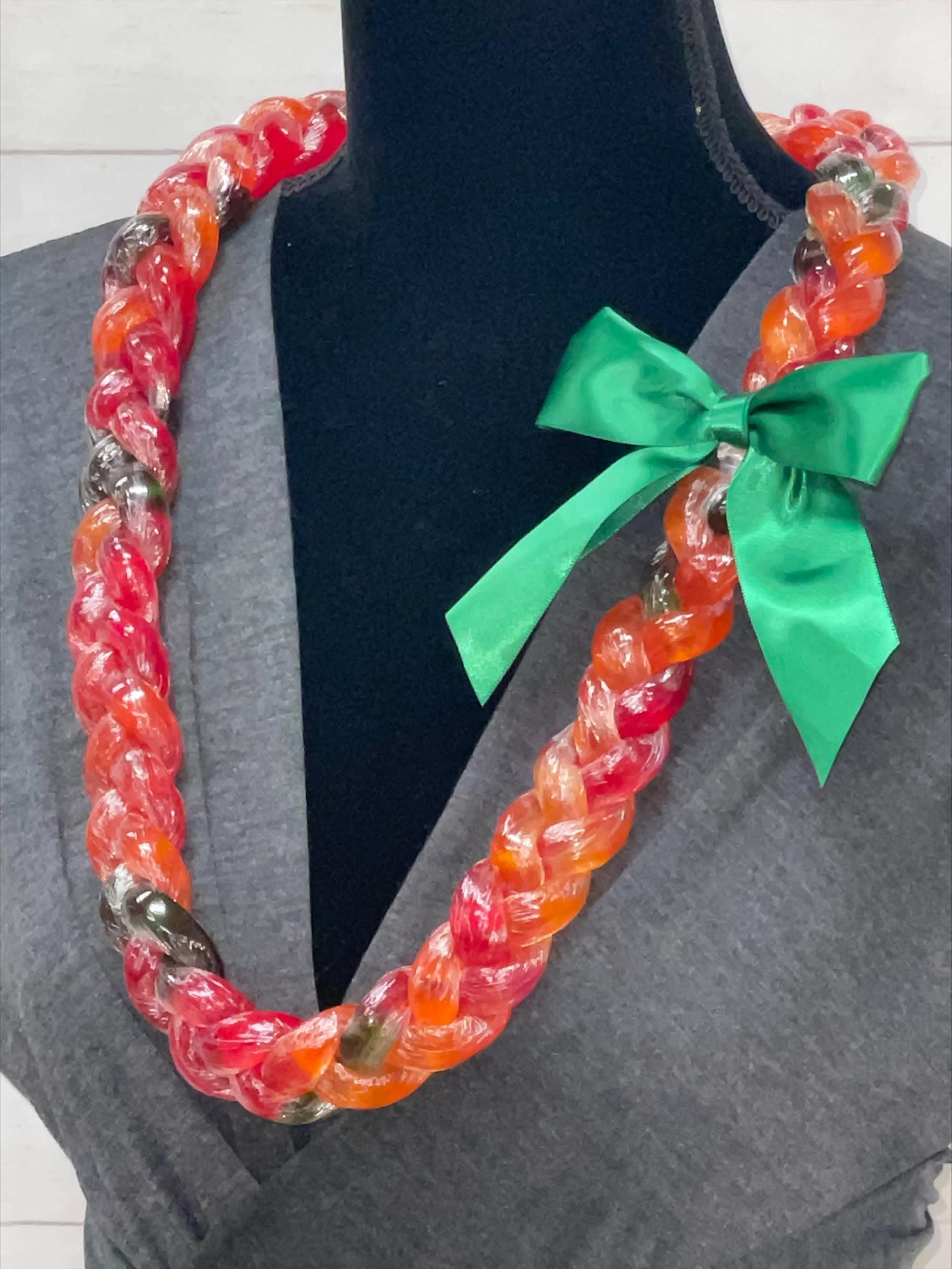 Braided candy lei in red and orange hues with a green satin bow on a display bust.