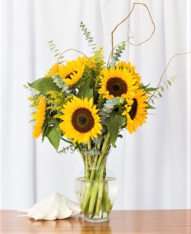 Tall bouquet of bright yellow sunflowers in a clear glass vase on a wooden surface.