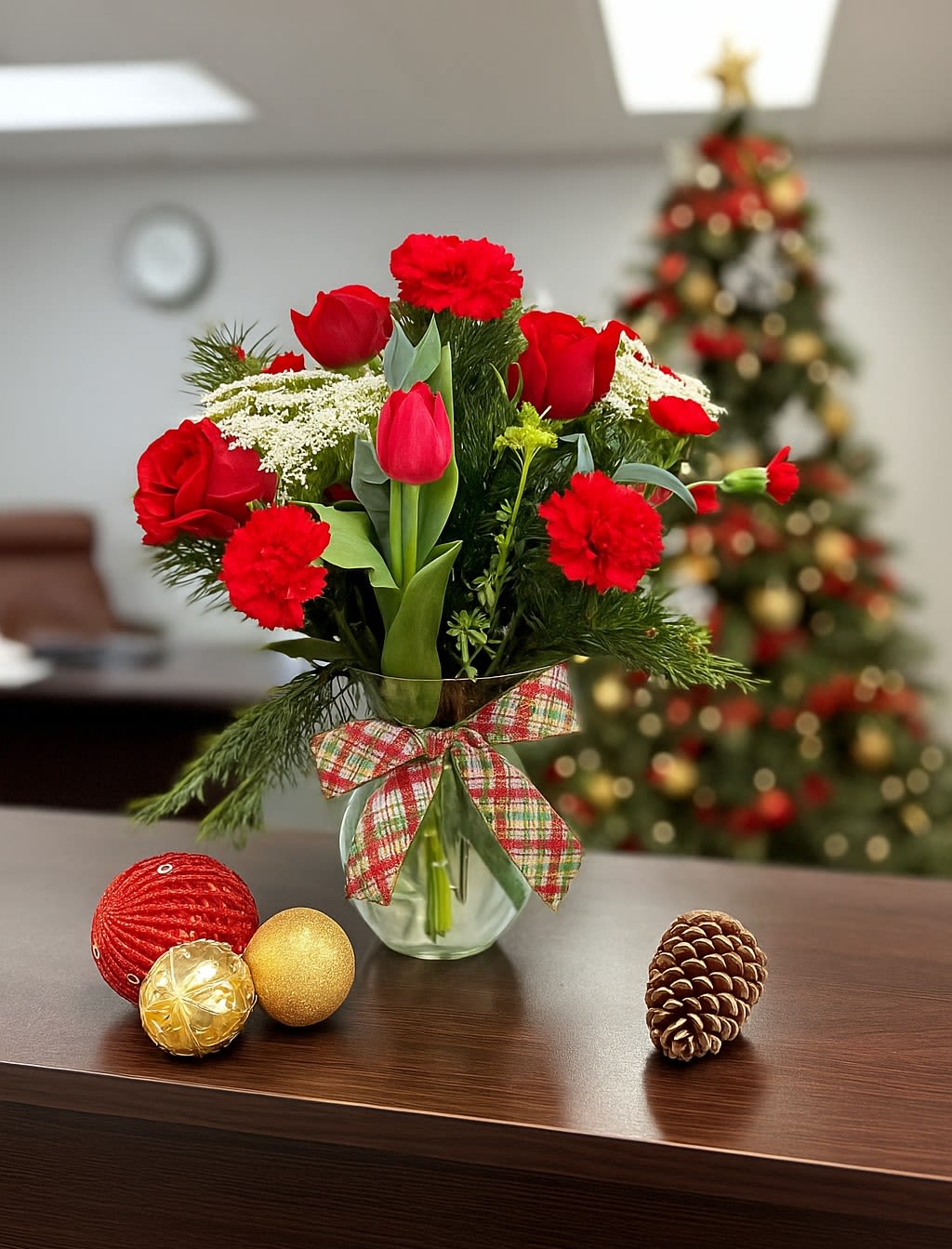 Red carnations and tulips in a glass vase with plaid bow, with ornaments and a pinecone on the table.