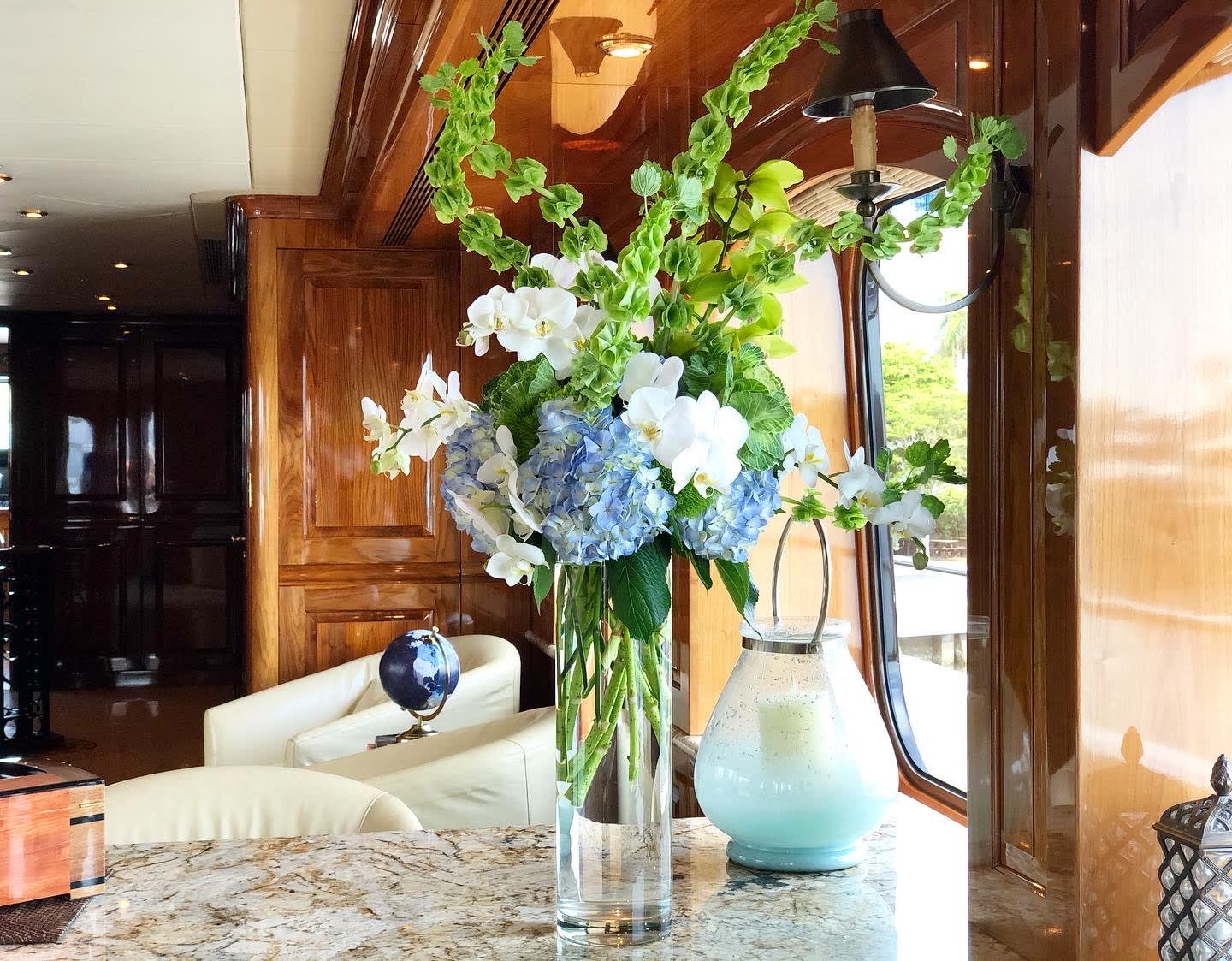 Tall floral arrangement with blue hydrangeas, white orchids, and green hanging garlands in a glass vase