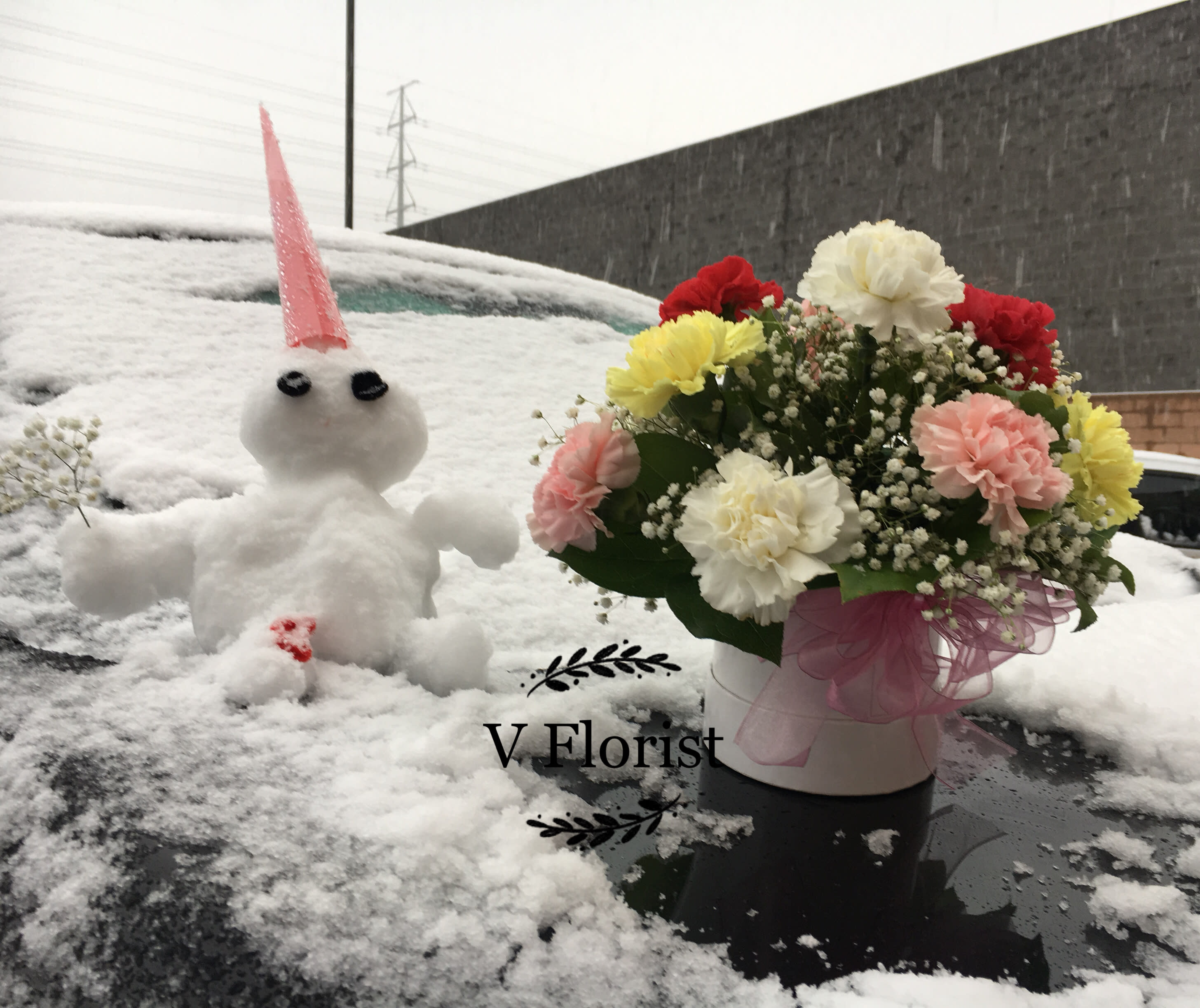Round white box of mixed carnations with baby’s breath in snow beside a small snow figure.