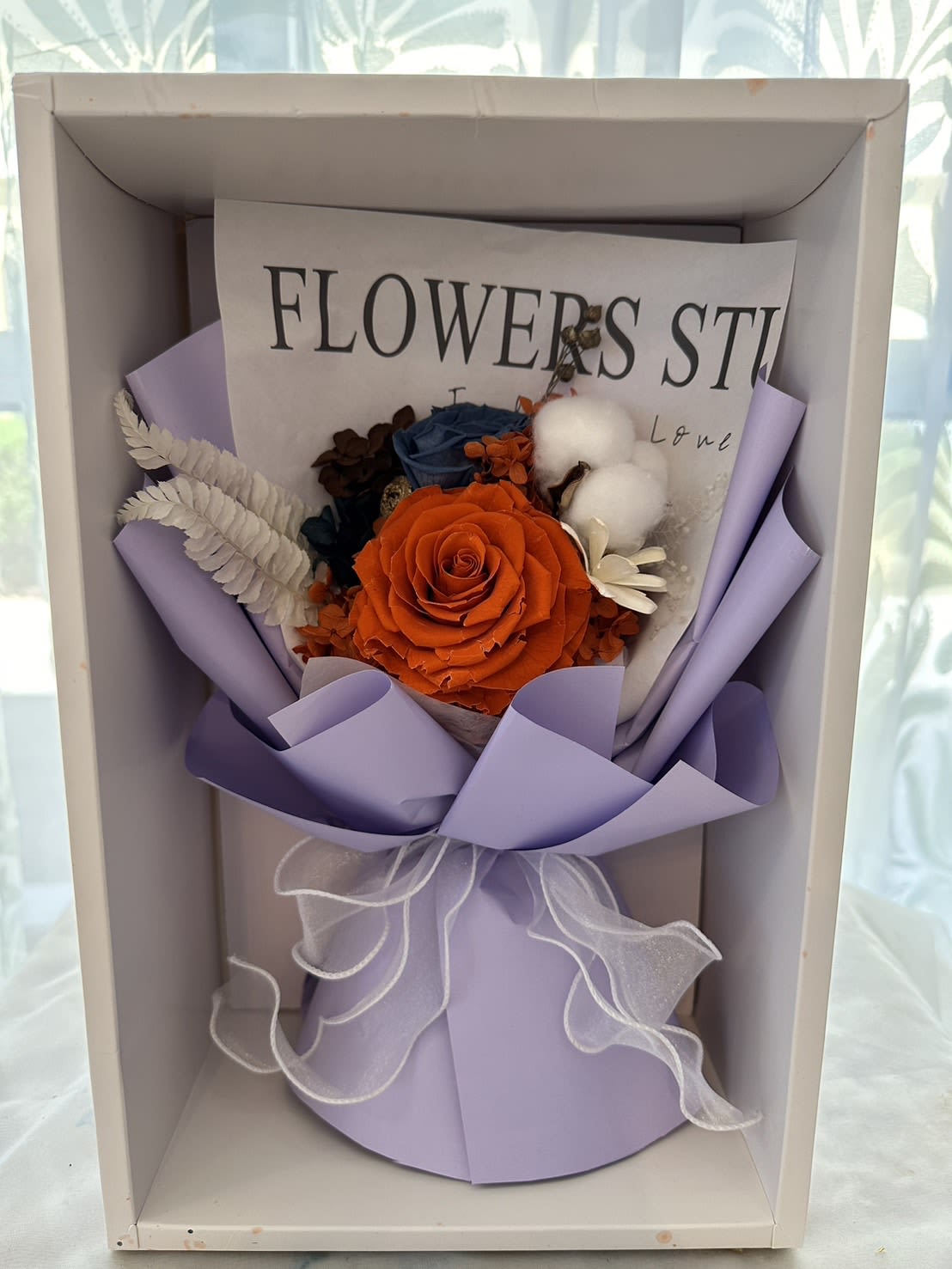 Boxed arrangement with orange rose, blue blooms, and cotton in lavender wrap with white ribbon