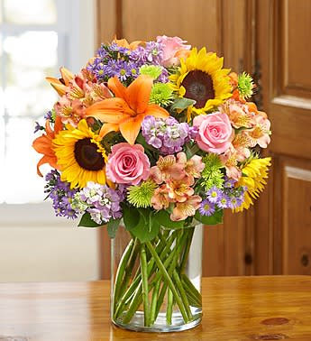 Mixed bouquet of sunflowers, roses, lilies, and purple flowers in a glass vase