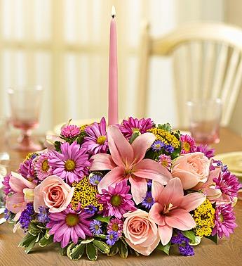 Low floral centerpiece with pink lilies, roses, and purple daisies around a tall pink candle.