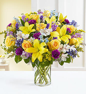 Bright mixed bouquet of yellow lilies, roses, and purple flowers in a glass vase