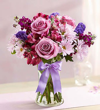 Pink roses and purple flowers in a clear glass vase with a lavender ribbon