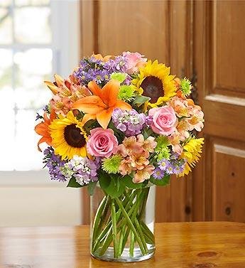 Mixed bouquet of sunflowers, roses, lilies, and purple flowers in a glass vase