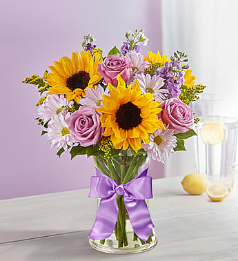 Bouquet of sunflowers, pink roses, and daisies in a glass vase with a purple ribbon