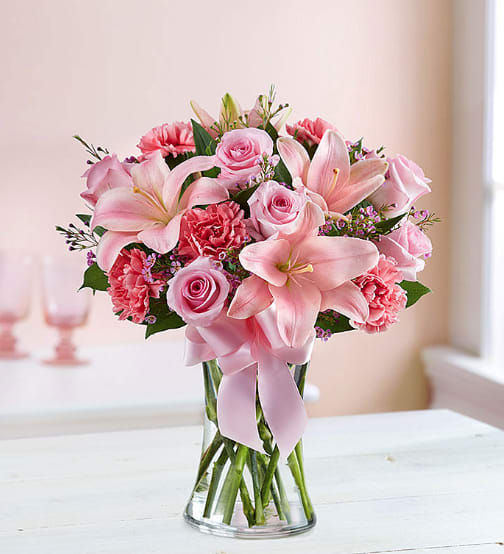 Pink lilies, roses, and carnations in a clear glass vase with a pink ribbon