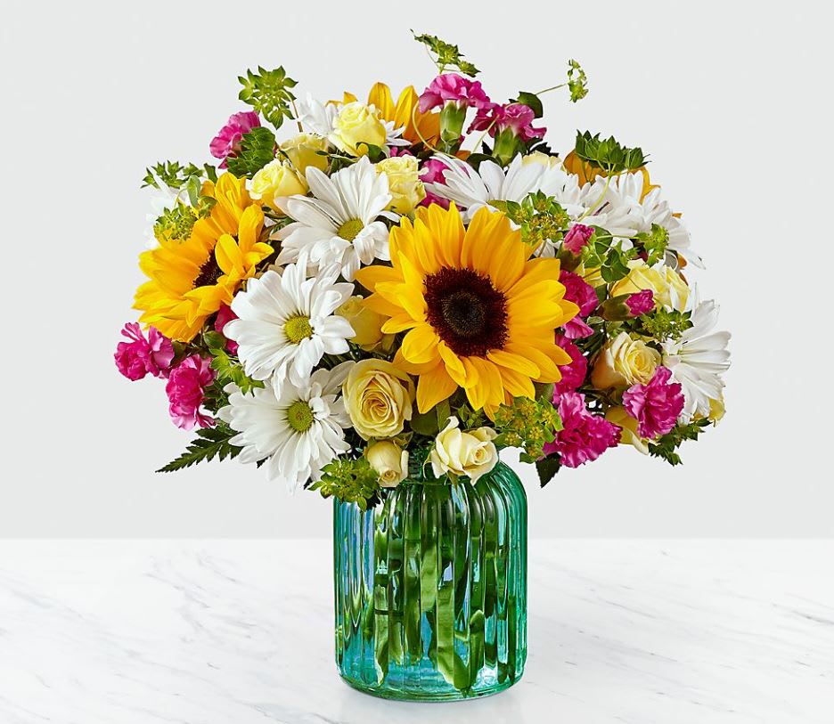 Sunlit Meadows™ Bouquet - Premium - With its sunny hues and fun floral variety, this bouquet will light up the face of whoever you give it to. Through a bright combination of sunflowers, daisy pompons, and spray roses set in a ribbed aqua glass vase, our FTD florists have designed a bouquet that brighten up every moment. GOOD bouquet is approx. 11"H x 11"W. BETTER bouquet is approx. 12"H x 12"W. BEST bouquet is approx. 13"H x 13"W. EXQUISITE bouquet is approx. 14"H x 13"W.  