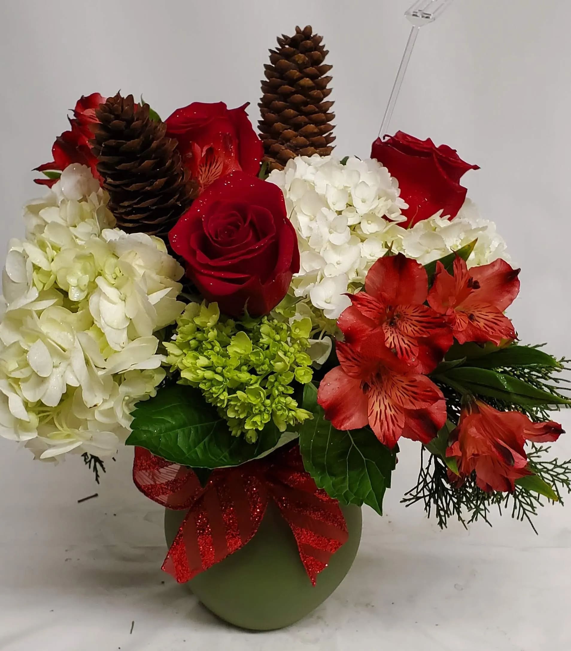 Red roses and white hydrangeas in a green vase with pinecones and a red ribbon