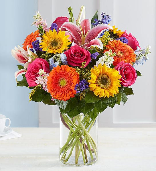 Mixed bouquet of roses, lilies, sunflowers, and gerbera daisies in a glass vase