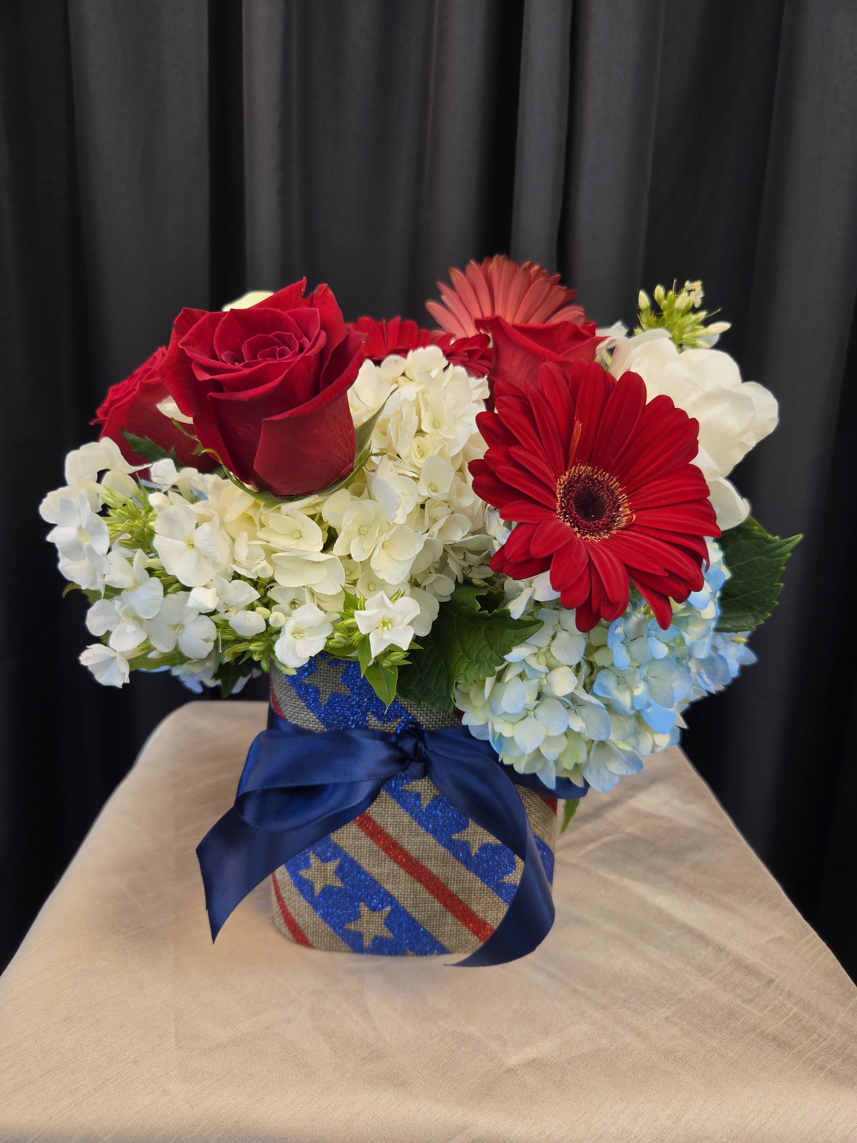 CLASSIC AMERICANA - Classic Americana is a vibrant fresh flower arrangement that celebrates timeless American pride with bold color and elegant style. Designed in a contemporary glass cube wrapped with patriotic ribbon, this handcrafted floral arrangement features lush white and blue hydrangea, vibrant red Gerbera daisies, and classic red and white roses. The striking combination of red, white, and blue blooms creates a cheerful and sophisticated display, perfect for Memorial Day, Independence Day, Veterans Day, military homecomings, or honoring someone special. Handcrafted by our expert florists, Classic Americana is a meaningful floral gift that brings beauty, gratitude, and patriotic spirit to any home, office, or celebration.